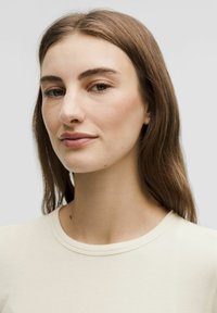 Helle beige gerippte T-Shirt mit rundem Halsausschnitt. Glatte Textur und taillierte Passform. Modell mit langen braunen Haaren und minimalem Schmuck.
