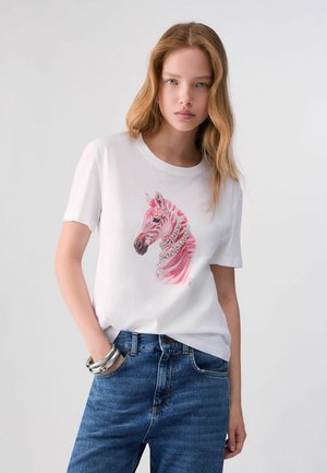 Jeune femme aux longs cheveux blonds porte un t-shirt blanc à motif zèbre rose, un jean bleu et des bracelets argentés, debout devant un fond uni.
