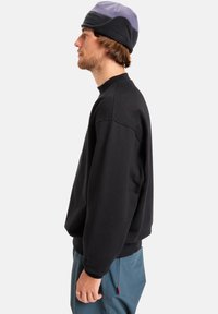Zwarte sweatshirt met een ronde hals, met lange mouwen en een relaxte pasvorm. Draagt bij lichtblauwe broek en een veelkleurige muts.