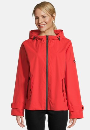 Femme souriante portant une veste à capuche rouge vif avec zipper frontal, poches à rabat et poignets réglables, sur un fond clair uni.