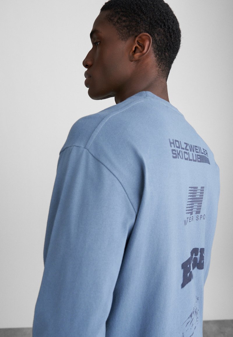Világoskék pulóveres sweatshirt. Sötétkék szöveget tartalmazó grafika található a hátsó részen, bordázott mandzsettákkal és klasszikus kerek nyakú kialakítással.
