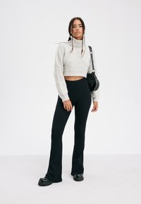 Lichtgrijze cropped trui met ribbelstructuur, zwarte high-waisted flare broek en stevige zwarte plateau schoenen; houdt een zwarte handtas vast.