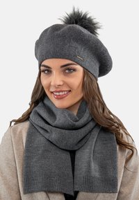 Béret en laine grise avec un pompon en fausse fourrure, associé à une écharpe grise assortie avec des extrémités côtelées et une texture lisse.