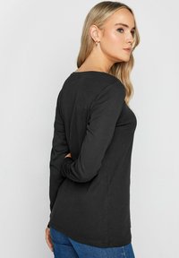 Schwarzes Langarmoberteil aus weichem Material, mit einem runden Ausschnitt und lässiger Passform. Stylisch zu blauen Jeans getragen, von hinten betrachtet.