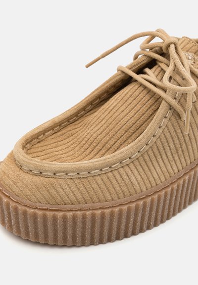 Pantofii slip-on din bumbac corduroy de culoare bej, cu un superior texturat, cusături albe de-a lungul gulerului și o talpă din cauciuc nervurată pentru aderență și confort.