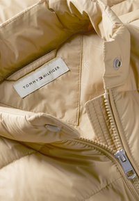 Veste matelassée beige avec une étiquette Tommy Hilfiger à l'intérieur près du col et une fermeture éclair métallique partiellement fermée à l'avant.