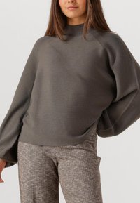 Grau gestrickter Pullover mit Stehkragen, lässiger Silhouette und weiten Ärmeln, kombiniert mit braunen Hahnentritt-Hosen.