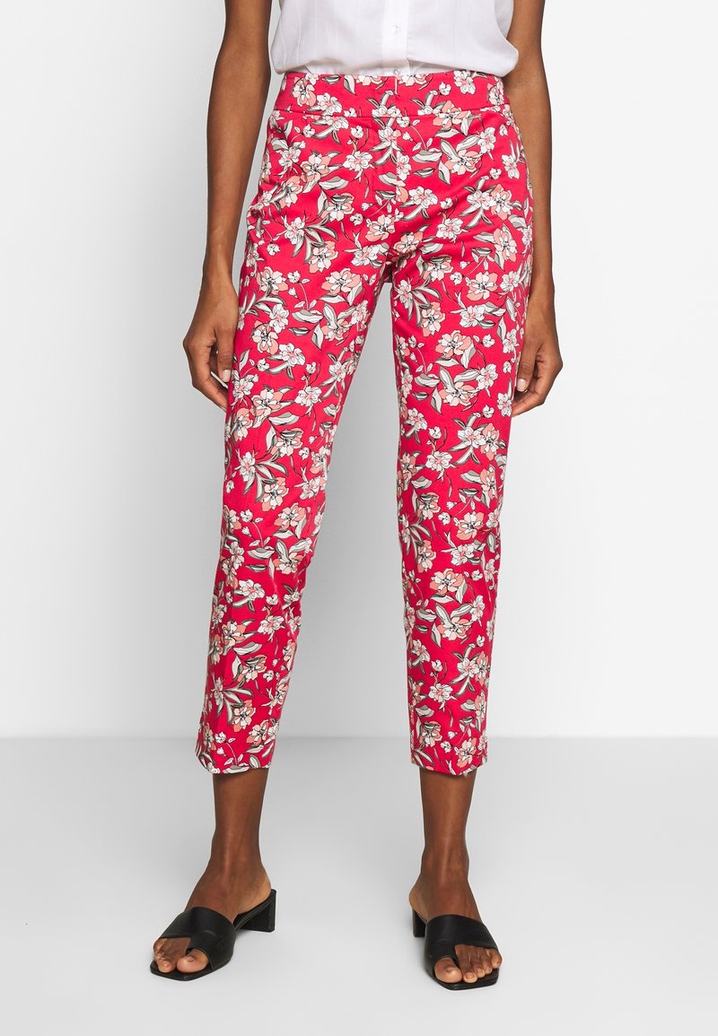 Femme portant un pantalon rouge court avec un imprimé floral blanc et rose, associé à un chemisier blanc et des sandales noires à talons ouvertes.