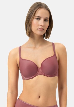 Junge Frau mit kurzen hellbraunen Haaren, die einen mauve gepolsterten Bügel-BH und passende Unterteile vor einem schlichten Hintergrund trägt.