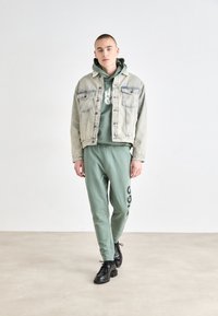 Chaqueta de mezclilla gris claro sobre un hoodie verde y joggers verdes a juego con un logo "HUGO". Unos zapatos negros de cordones completan el conjunto.