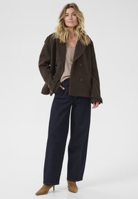 Une femme se tenant debout, vêtue d'un manteau marron croisé, d'un top beige, de jeans larges foncés et de bottines à talons beiges, sur un fond uni.