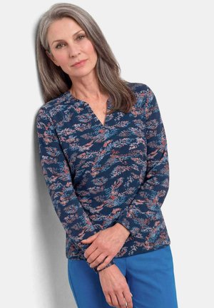 Vrouw met grijs haar, gekleed in een blauwe bloemenprint blouse met lange mouwen en een blauwe broek, staand met gevouwen handen tegen een lichte achtergrond.