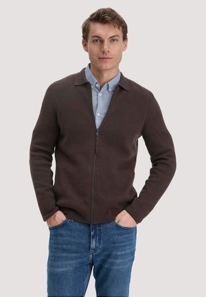 Junger Mann, der einen braunen Zip-Up-Pullover über einem hellblauen Hemd mit Knöpfen und blauen Jeans trägt, steht mit den Händen in den Taschen.