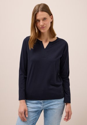 Chemise à manches longues navy avec un col en V et des détails de couture discrets sur la poitrine. Fabriquée dans un tissu doux et léger. Assortie avec un jean clair.
