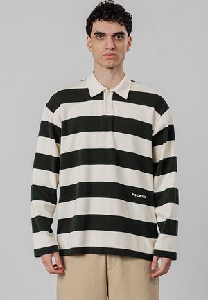 Ung mand med kort krøllet hår iført en sort-hvid langærmet polo med vandrette striber og beige bukser mod en ensfarvet baggrund.