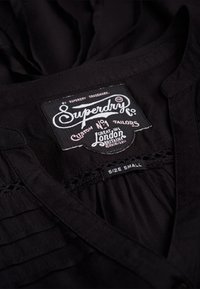 Chemise noire avec une étiquette affichant "Superdry Co," "Custom No. 1 Tailors," et "Taille S." La chemise présente des détails plissés près du col.