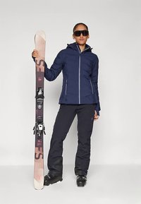 Osoba nosící námořnicky modrou lyžařskou bundu a černé lyžařské kalhoty drží svisle pár lyží, na očích má velké černé lyžařské brýle proti jednoduchému pozadí.