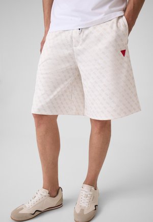 Man wearing white patterned shorts with hands in pockets, white sneakers, and a white shirt, standing against a plain light background.