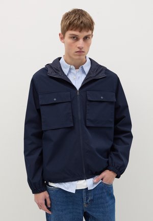 Chaqueta con capucha en color marino, con cierre en la parte delantera, dos bolsillos en el pecho, puños elásticos y un corte relajado. Lleva una camisa de rayas en azul claro debajo.
