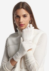 Weiße Handschuhe aus weichem Strickmaterial mit einem gerippten Handgelenk. Das Modell trägt eine weiße ZIP-Weste und zeigt eine neutrale Farbpalette.