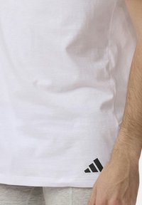 Biała sportowa koszulka z małym czarnym logo Adidasa blisko dołu oraz widoczna ręka osoby po prawej stronie.