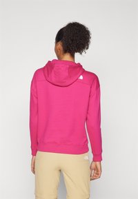 Felpa con cappuccio rosa con polsini e orlo a costine, caratterizzata da un piccolo logo bianco sulla parte superiore della schiena. Indossata con pantaloni beige chiaro.
