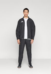 Μαύρο αθλητικό σετ από ελαφρύ ύφασμα, με ελαστική μέση, ζακέτα με φερμουάρ και λευκές λεπτομέρειες λογότυπου.