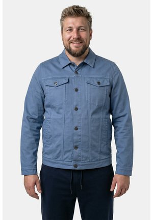 Lächelnder Mann mit kurzem blondem Haar und Bart trägt eine blaue Jacke mit Knöpfen und dunkle Hose, steht vor einem schlichten hellen Hintergrund.