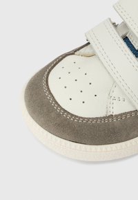 Λευκό δερμάτινο και γκρι σουέτ αθλητικό παπούτσι με διάτρητη μύτη, ρυθμιζόμενο λουράκι και υφή στη σόλα. Ορατή ραφή κατά μήκος των άκρων.