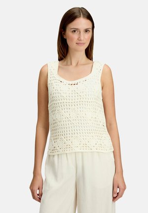 Vrouw draagt een gebroken wit gehaakt mouwloos topje met bloemmotieven en losse gebroken witte broek, staand tegen een effen achtergrond.