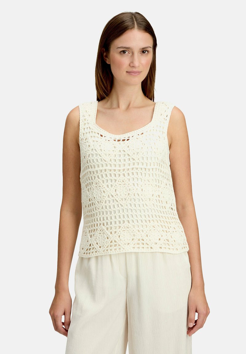 Femme portant un haut sans manches en crochet blanc cassé avec des motifs floraux et un pantalon ample blanc cassé, debout devant un fond uni.