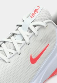 Ανοιχτό γκρι αθλητικό παπούτσι με διχτυωτό άνω μέρος, λευκές κορδόνες και φωτεινό ροζ σήμα της Nike. Υφή επιφάνειας, με επενδεδυμένο πάτο σε αντίθεση λευκού και ροζ.