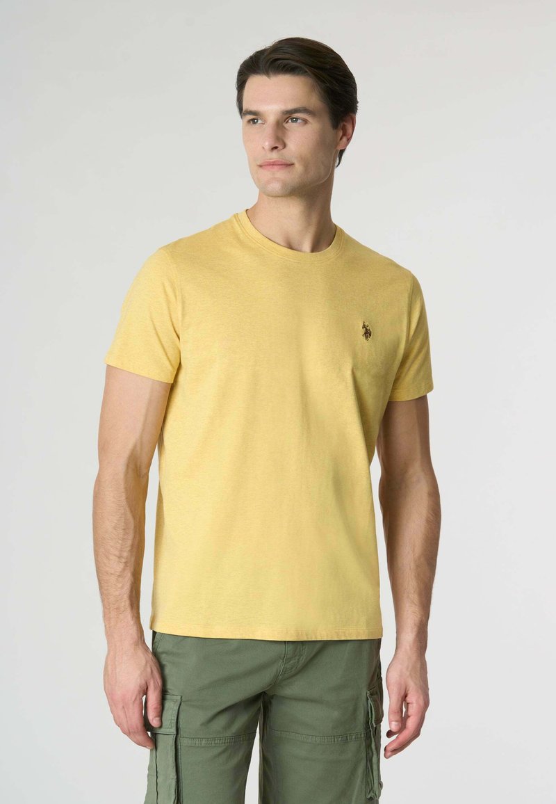 T-shirt di cotone gialla con collo rotondo classico e maniche corte, caratterizzata da un piccolo logo scuro sul lato sinistro del petto. Abbinata a pantaloni cargo verdi.