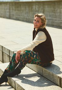 Bruine faux bont vest over een crèmekleurige coltrui, gecombineerd met gestreepte groene en oranje broeken. Zwarte hoge enkellaarsjes. Zittend op stenen treden.