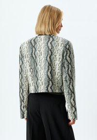 Veste à imprimé serpent grise et crème, coupée, avec un motif texturé. Manches longues et coupe ajustée, associée à un pantalon noir.