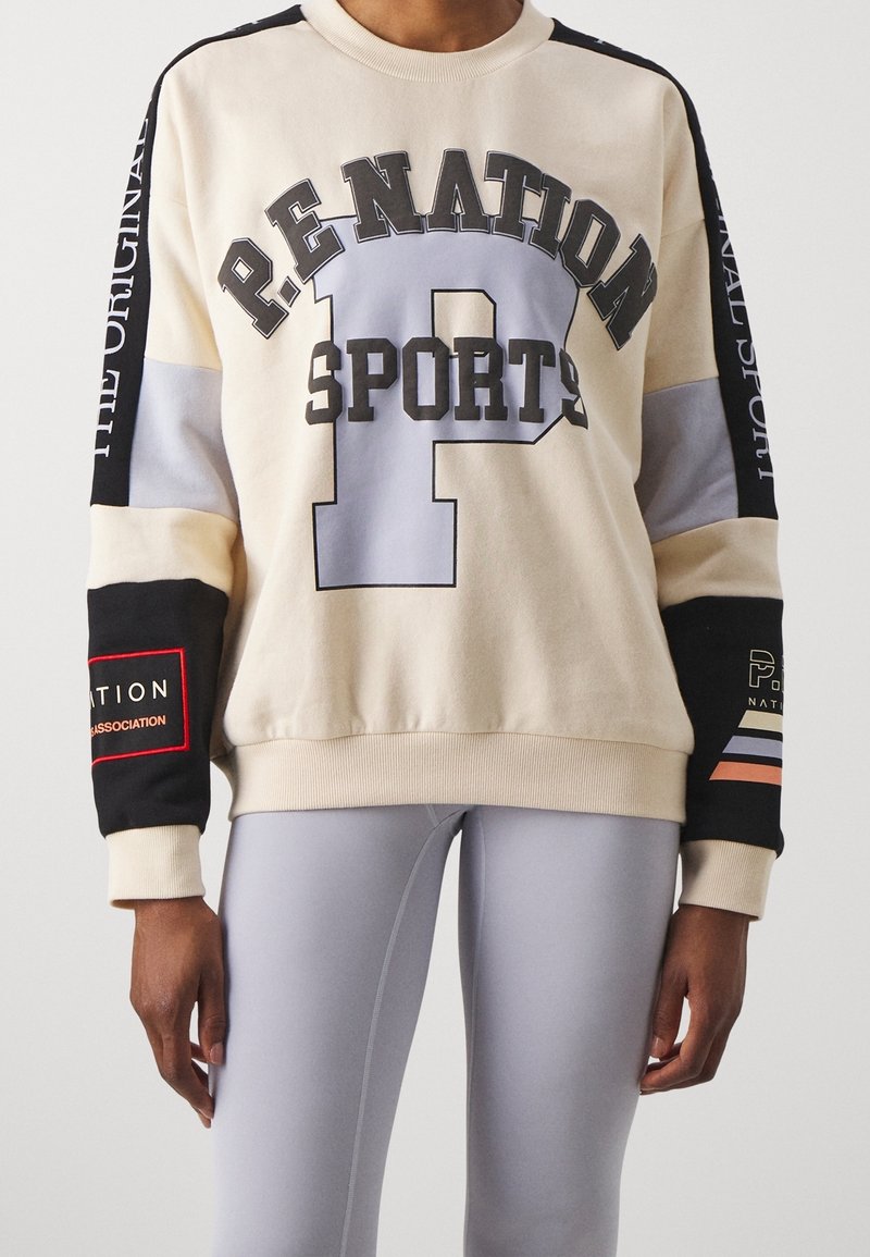 Personne portant un sweat-shirt beige avec le texte "P.E NATION SPORTS" et des détails en blocs de couleur noir, gris et orange sur les manches, ainsi que des leggings gris clair.