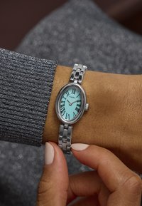 Hõbedane roostevabast terasest kell ovaalse korpusega, akvamariinist kelladial, mustade Rooma numbritega ja ahelkettiga, kantud käe peal.