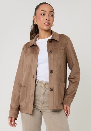 Kobieta ubrana w jasnobrązową zamszową kurtkę na białej koszuli i beżowych spodniach, stojąca na tle jednolitego szarego tła.