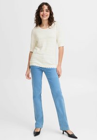 Ljust krämfärgad, texturerad stickad topp med scalloperad ringning och fåll, kombinerad med ljusblå raka jeans och svarta högklackade skor.