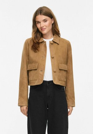 Vrouw draagt een beige suède cropped jas met knopen en klepzakken, witte top en zwarte broek met hoge taille, staand tegen een witte achtergrond.