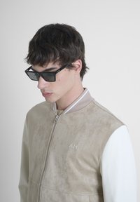 Chaqueta bomber de ante beige con mangas de cuero blanco, cierre frontal de cremallera, cuello acanalado y logotipo bordado. Gafas de sol rectangulares negras.