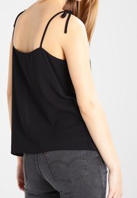 Débardeur noir sans manches avec des bretelles réglables, fabriqué dans un tissu léger. Il présente une coupe décontractée et une texture lisse, associé à un jean gris.