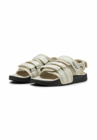Beige sandaler med justerbare Velcro-remme, gummisål og blød fodseng. Har vandrette striber og branding på siden.