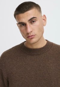 Brūns adīts džemperis ar ribotu apkakli, mīkstu tekstūru un vidēji smagu materiālu. Tuva piegriezuma dizains ar minimāliem šuvju detalizējumiem.