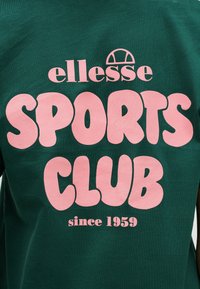 Πράσινο βαμβακερό μπλουζάκι με μεγάλο ροζ κείμενο που γράφει «ellesse SPORTS CLUB since 1959.» Στρογγυλεμένα γράμματα και ομαλή υφή.
