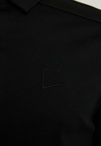Camisa de algodón negra con cuello puntiagudo y un sutil bordado de cuadrados negros en el frente, con una textura suave y sin patrones.