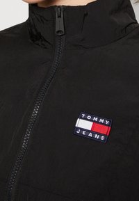 Czarna kurtka zapinana na zamek, wykonana z lekkiego materiału, z wysokim kołnierzem i małym haftowanym logo "TOMMY JEANS" w kolorach czerwonym, białym i niebieskim.