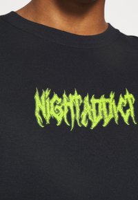 Black cotton t-shirt featuring bright neon green embroidered text "NIGHTADDICT" in a jagged, stylized font. Close-up view of neckline and fabric texture.