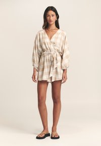 Šviesiai smėlio spalvos languotas kombinezonas su gilia V formos iškirpte, suspausta liemens dalimi ir balioninėmis rankovėmis, derinamas su paprastomis rudomis sandalais.