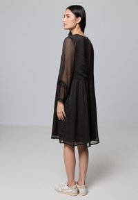 Vestido negro con mangas largas transparentes, diseño en capas, tela texturizada y dobladillo acampanado. Combinado con zapatillas blancas. Silueta simple y fluida.
